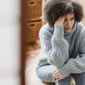 Young ethnic female in casual clothes sitting on pouf while crying with hand on head in light flat